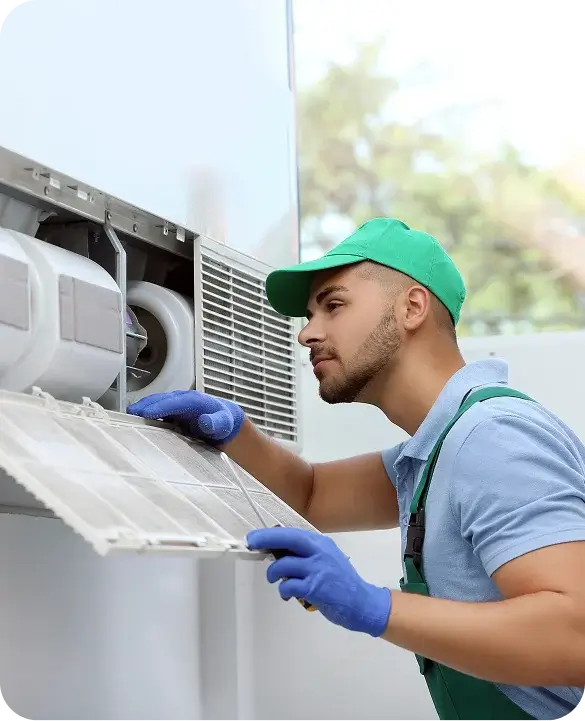 An HVAC technician in a green uniform and blue gloves carefully servicing an outdoor air conditioning unit with attention to detail.