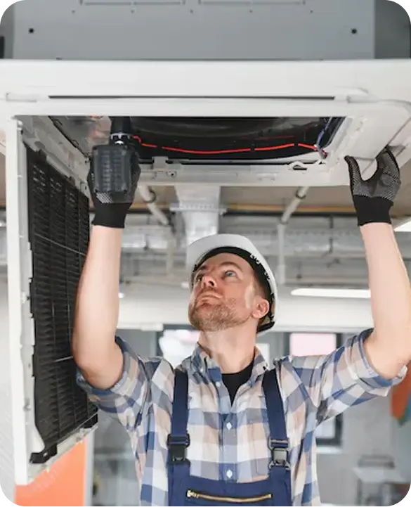 A technician in a hard hat and overalls installing a ceiling-mounted commercial HVAC unit in a commercial building.
