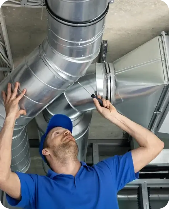 HVAC technician in blue uniform installing and repairing large silver metal ductwork on a commercial building ceiling