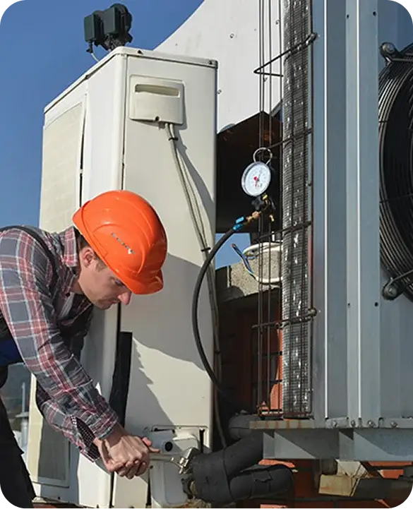 HVAC technician in orange hard hat servicing a large commercial rooftop air conditioning unit with pressure gauge attached